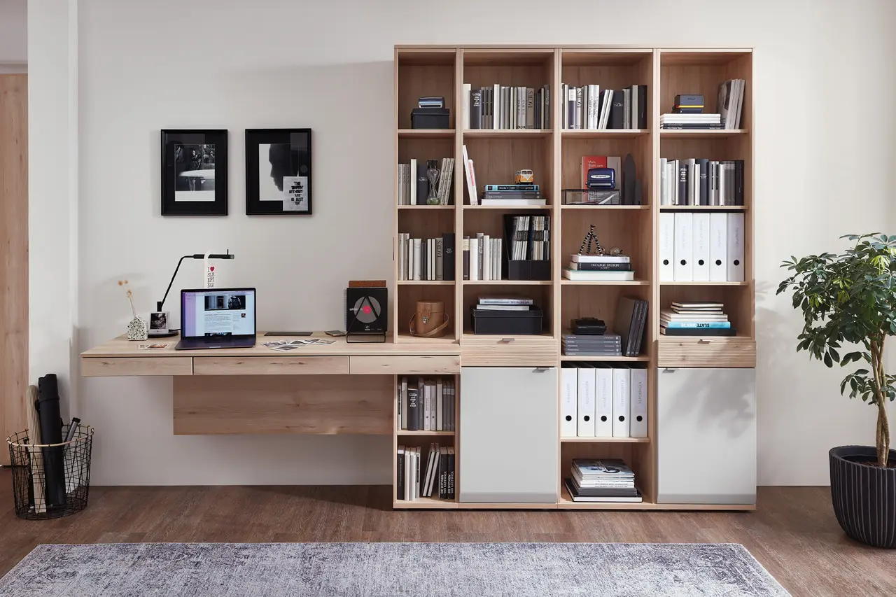 Schreibtisch aus Holz mit Laptop vor Regalwand mit Büchern und Ordnern, Bilder an der Wand und Pflanze rechts